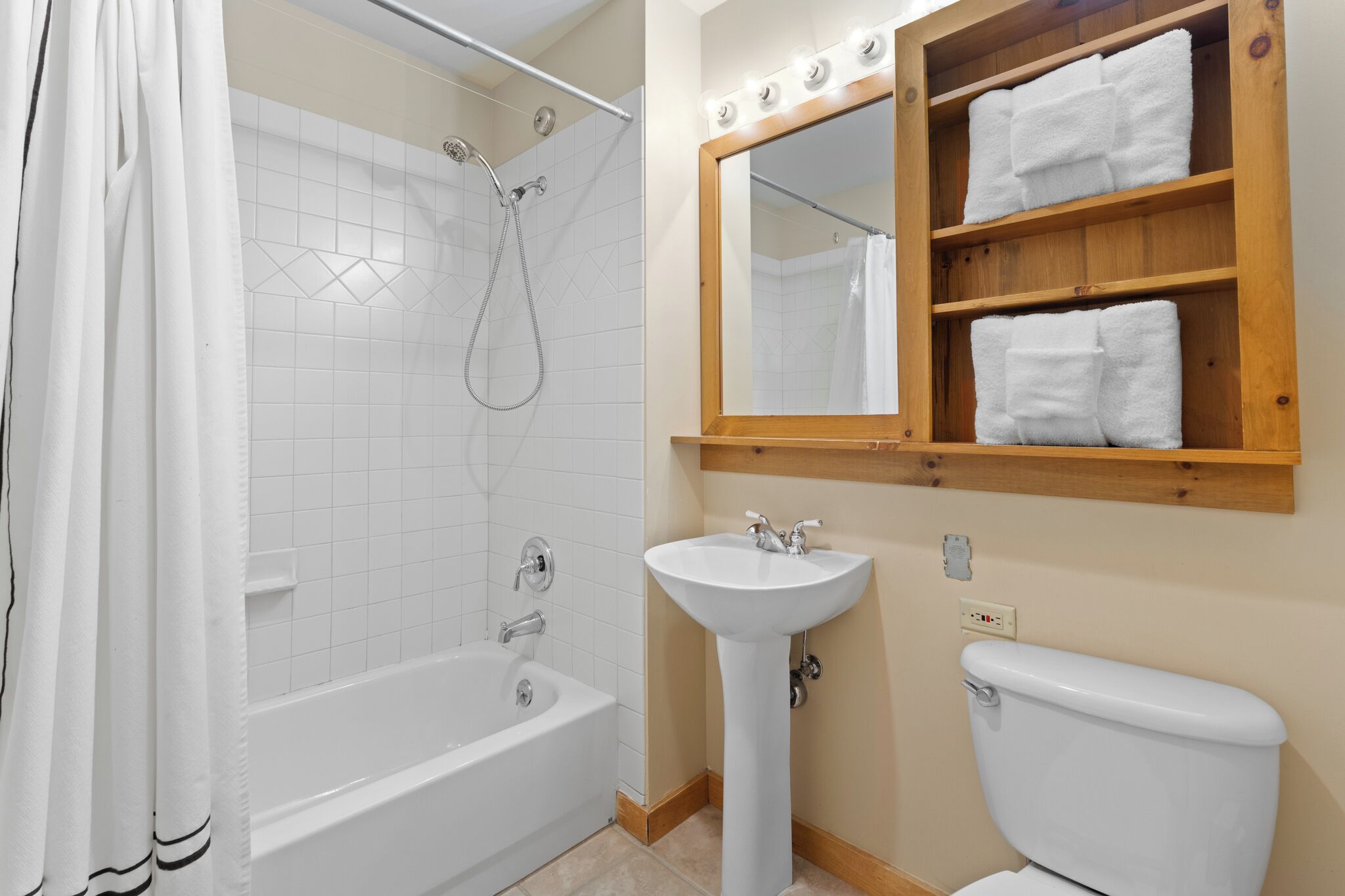 Une salle de bain avec une baignoire, un lavabo, des toilettes et une étagère en bois où sont posées des serviettes.