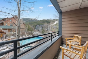 A balcony with a view of a swimming pool, wooden chairs, and a building with a sloped roof. - Dl8480-2 (Keystone)