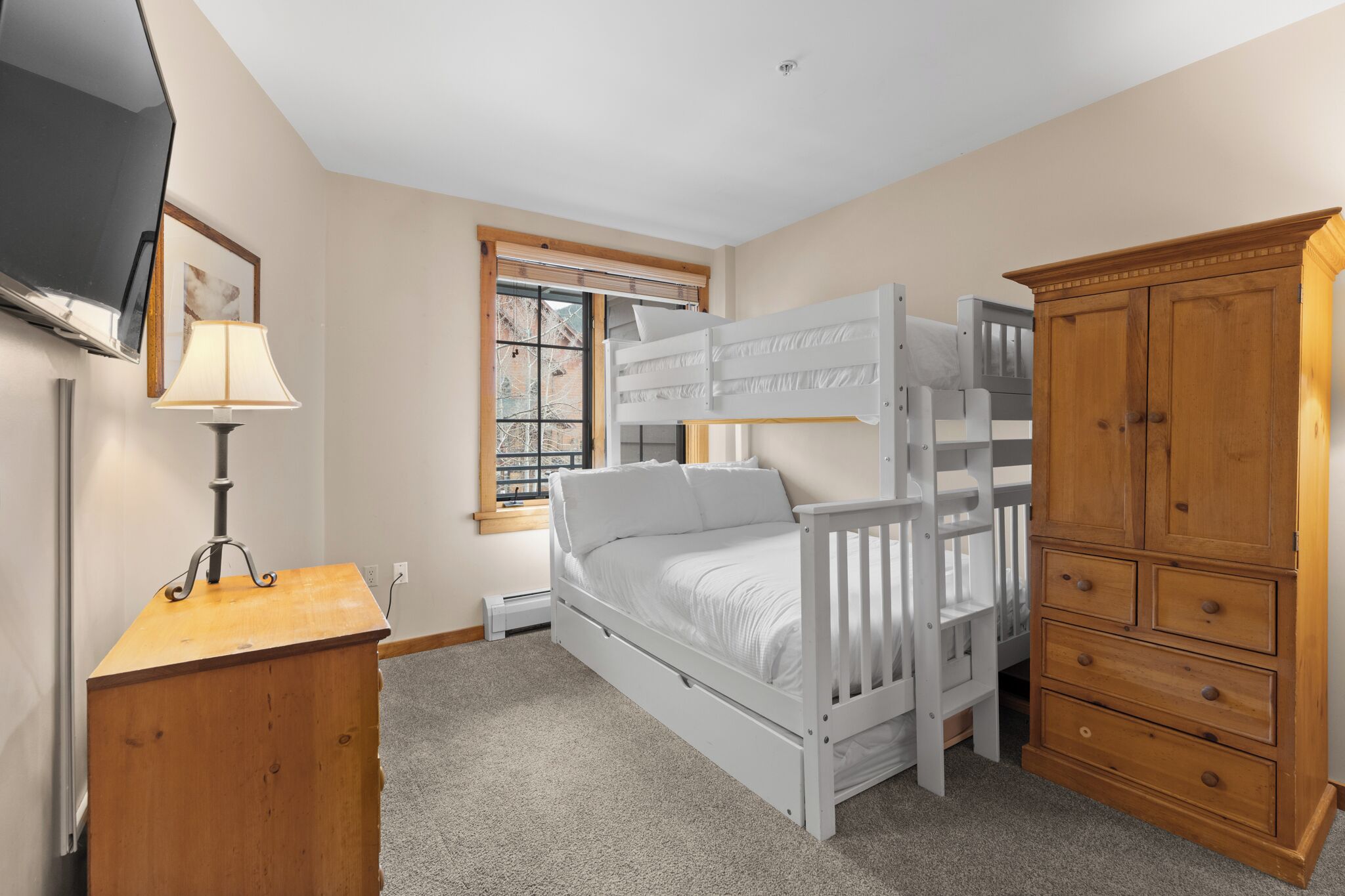 A bunk bed with a TV mounted on the wall, a wooden dresser, and a window with blinds.