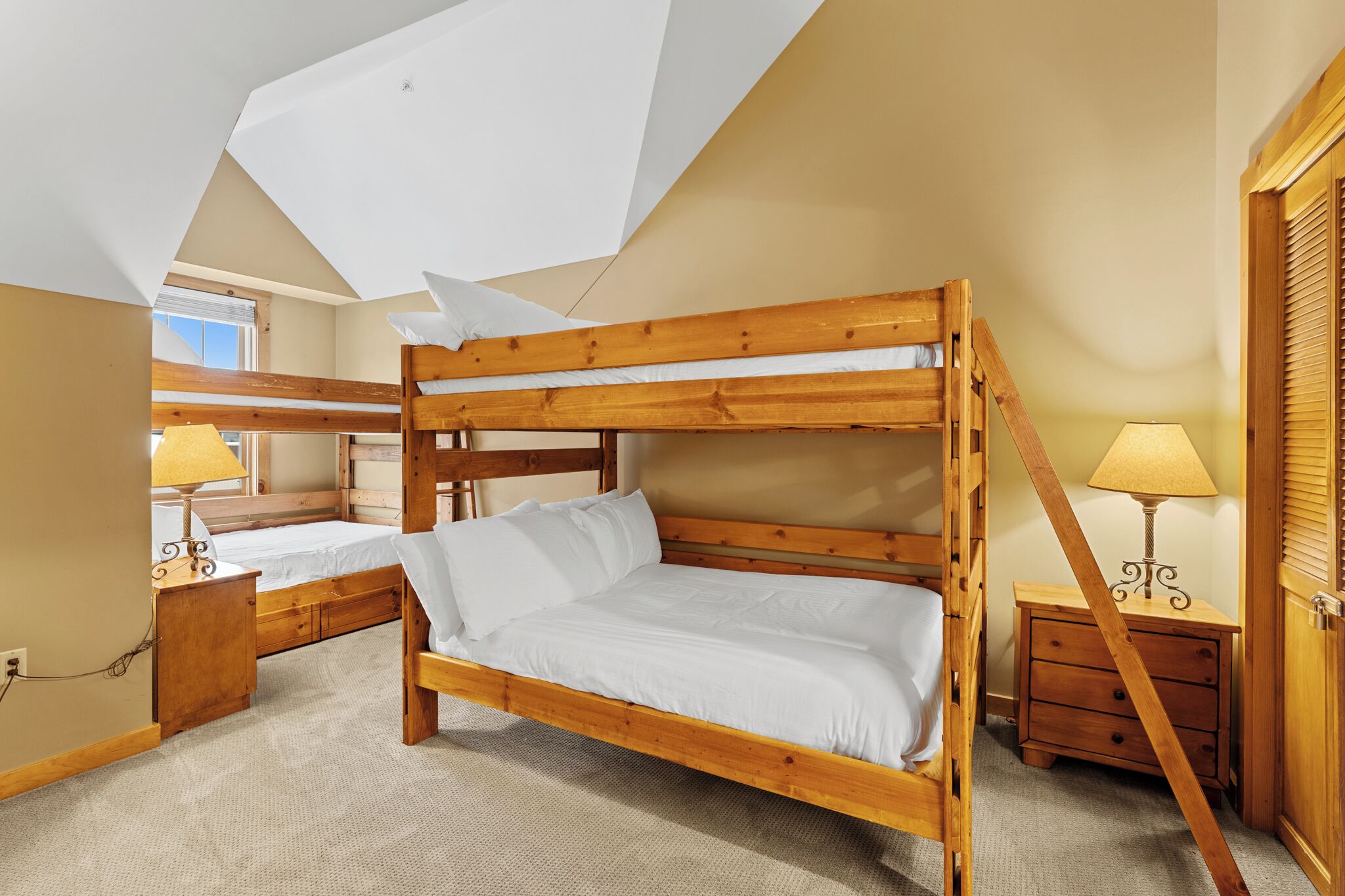 A bunk bed room with a wooden frame, a bedside table with a lamp, and a window with blinds.