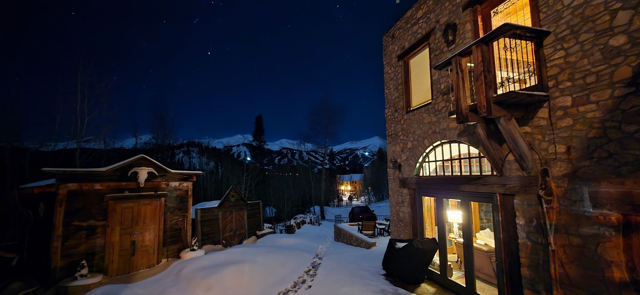 A snow-covered outdoor area with rustic buildings, a stone wall, and a wooden structure with a sloped roof.