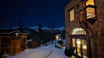 A snow-covered outdoor area with rustic buildings, a stone wall, and a wooden structure with a sloped roof.
