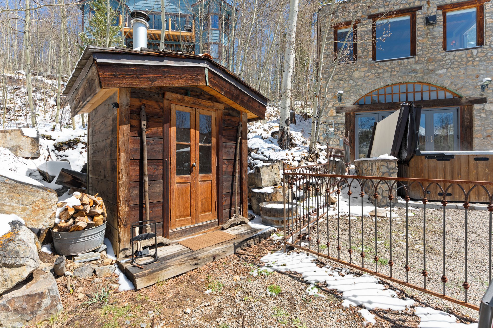Eine Holzhütte mit einem Metallzaun, ein Steinhaus im Hintergrund und ein Holzstapel.