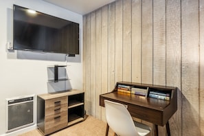 Ein Arbeitsraum mit einem Holztisch, einem Stuhl, einem an der Wand befestigten Fernseher und einem Bücherregal mit Büchern.