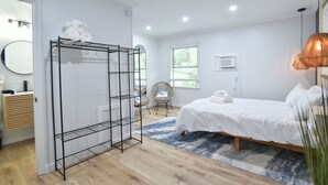 Quarto com cama, prateleira de metal preta com toalhas, cadeira de vime, tapete azul estampado e uma janela grande.