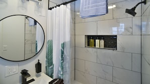 Casa de banho moderna com um espelho redondo, cortina de chuveiro, porta-toalhas e uma prateleira com artigos de higiene pessoal.