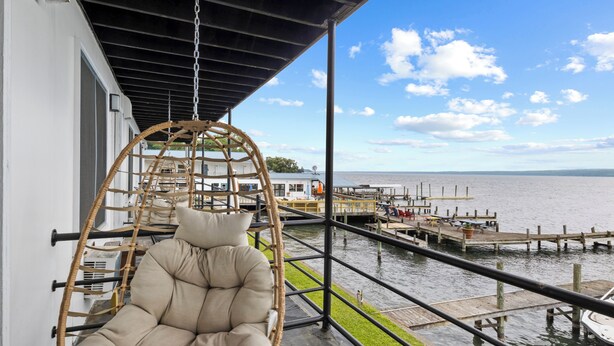A wicker swing chair on a balcony overlooking a waterfront with a dock and buildings in the background. - Double Room - 2 Queen Beds - Lake View (Himrod)