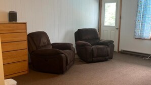 Une pièce avec deux fauteuils inclinables marron, une commode en bois, une porte blanche et une fenêtre avec des rideaux bleus.