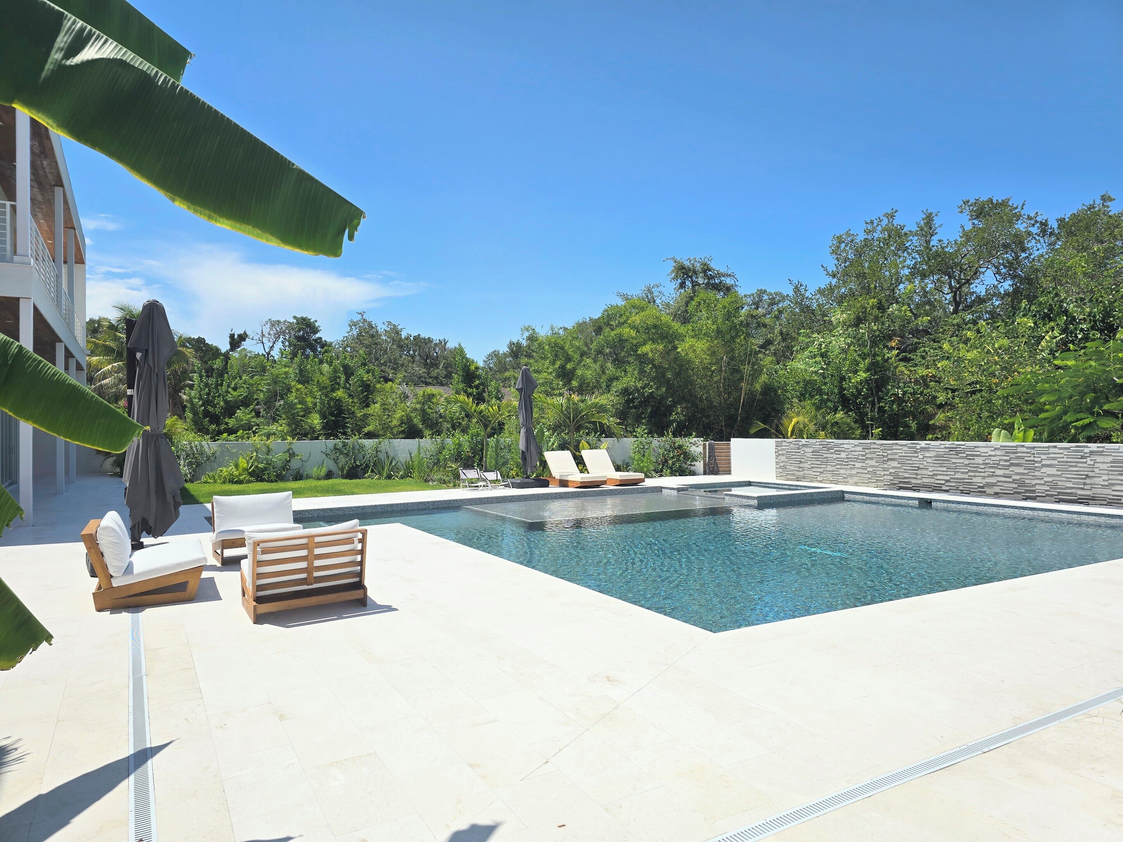 A rectangular swimming pool with lounge chairs and a covered umbrella, surrounded by a landscaped garden and a stone wall.