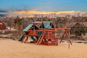 Une structure de jeux en bois, dotée de toboggans et de balançoires, se dressant sur fond de maisons et sous un ciel dégagé.