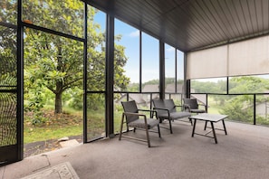 Un espace extérieur aménagé, abrité et doté de chaises et d’une table, de grandes fenêtres et d’une vue sur les arbres et les maisons.