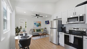 A modern kitchen with stainless steel appliances, a dining area with a white table and black chairs, and a living area with a gray sofa and colorful artwork. - Walk to Depot Town - Private Deck & King Bed (Ypsilanti)