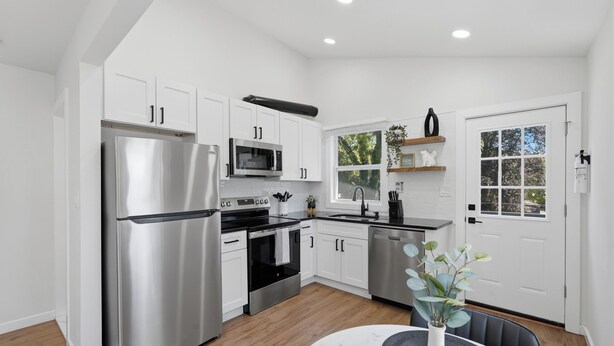A modern kitchen with stainless steel appliances, white cabinetry, and a dining area with a table and chairs. - Walk to Depot Town - Private Deck & King Bed (Ypsilanti)