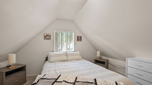 Une chambre avec un plafond en pente, un lit avec une literie blanche, deux tables de chevet avec des lampes, une commode et une fenêtre avec des stores.