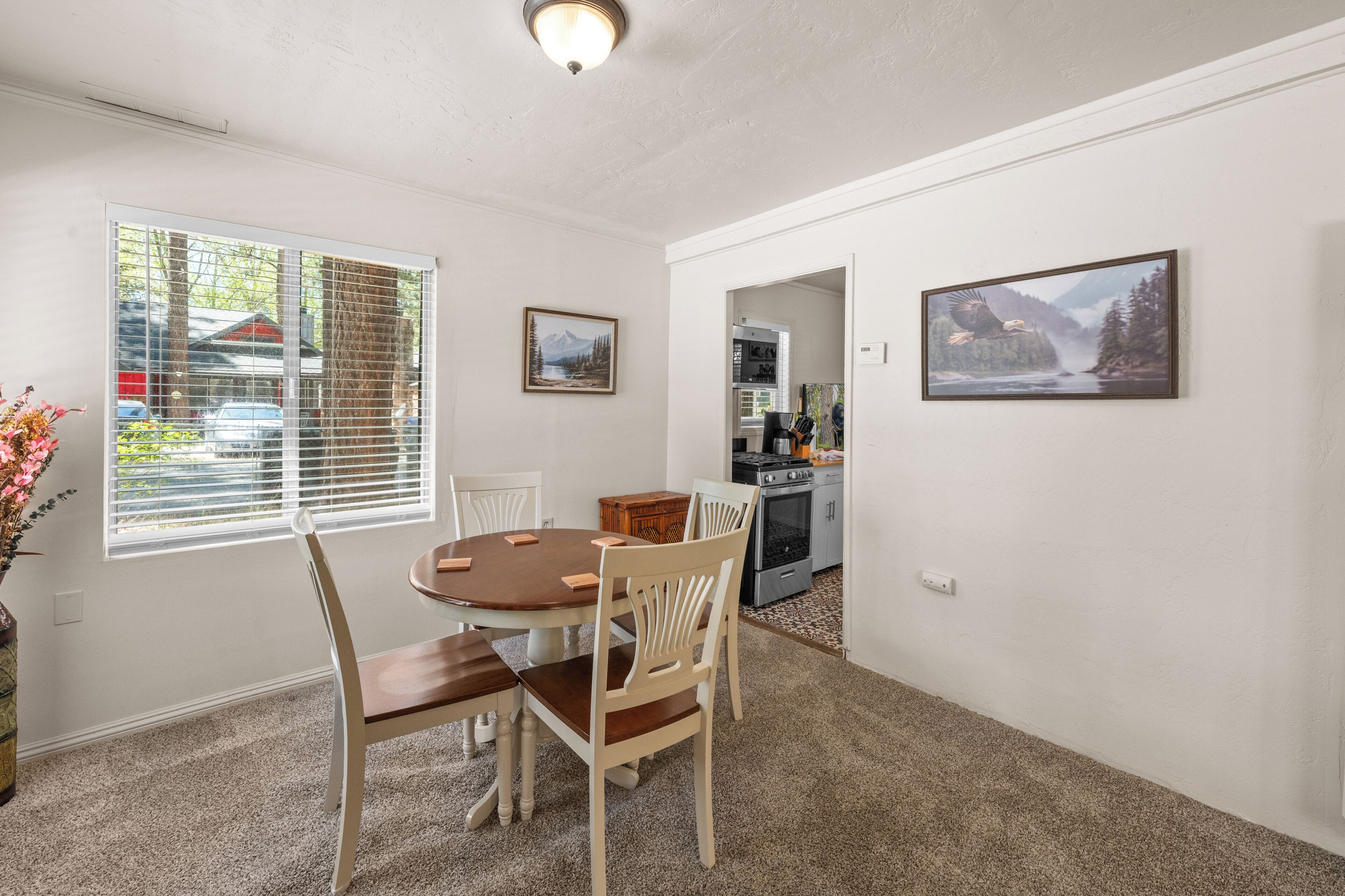A dining area with a round table, four chairs, a window with blinds, and two framed pictures on the wall.