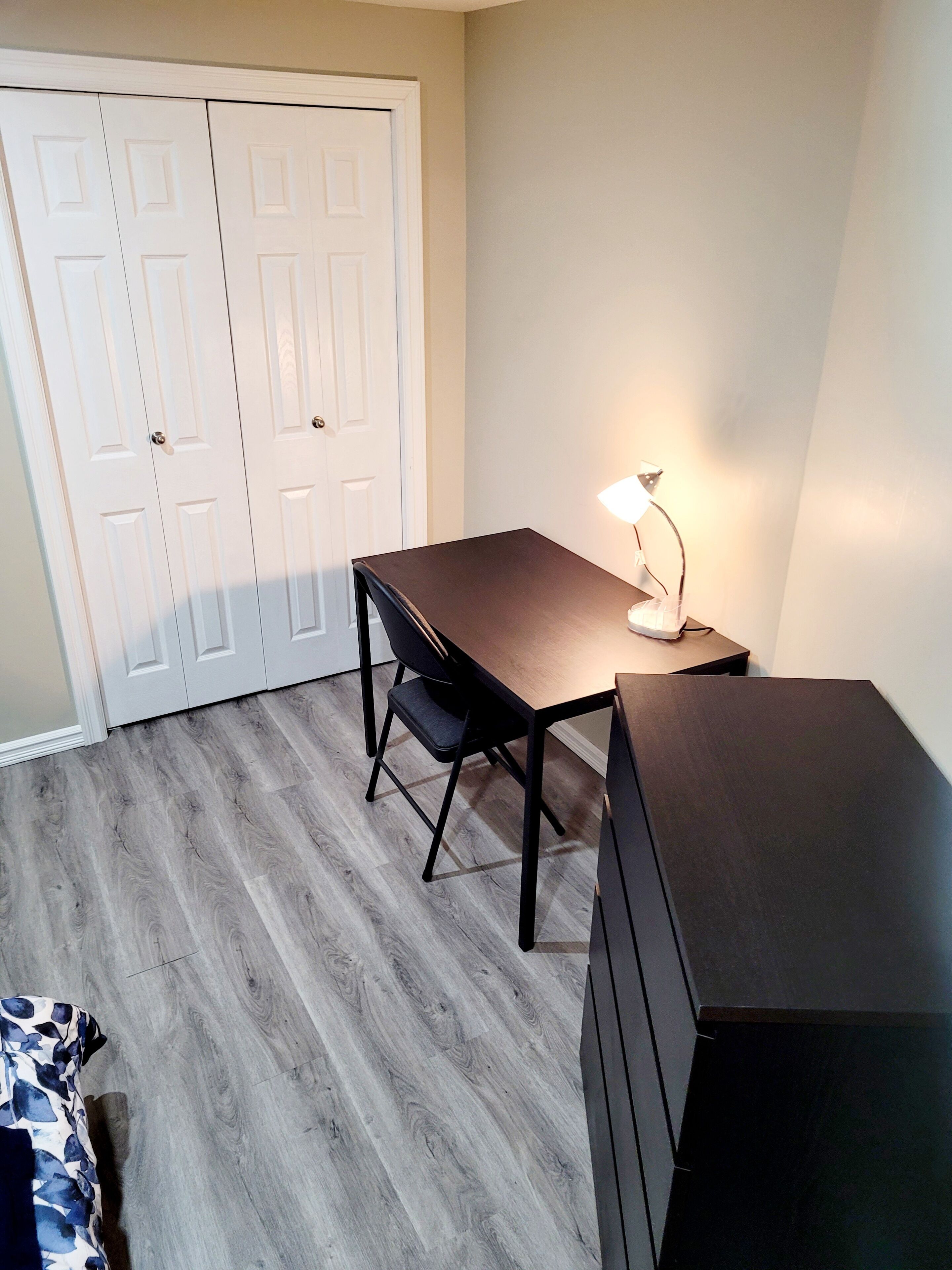 Una habitación con escritorio, silla y lámpara. Se observa un armario de dos puertas y una cama con colcha estampada.