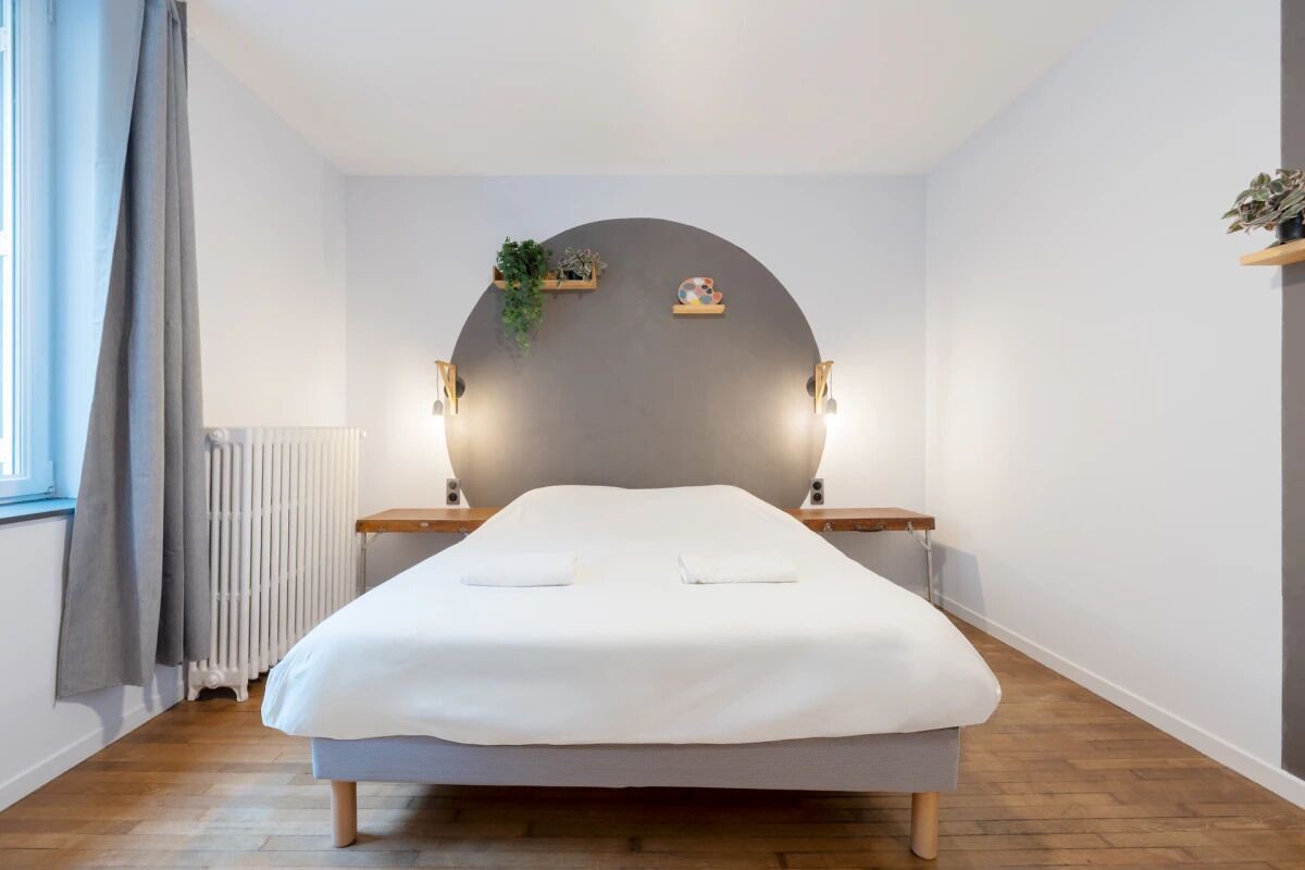 Une chambre à coucher au style minimaliste, avec un lit, des tables de chevet, un radiateur et une fenêtre avec des rideaux.