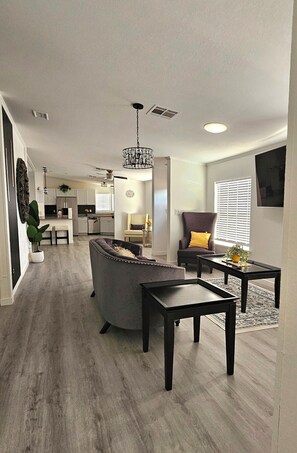 Un salon moderne avec un canapé, une table basse et un fauteuil, une cuisine en arrière-plan, et un coin repas avec une table et des chaises.
