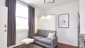 Ein modernes Wohnzimmer mit einem grauen Sofa, einem weißen, flauschigen Hocker, Holzboden, einer weißen Tür und einer Küchenzeile mit Herd.