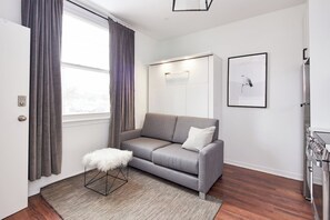 Un salon moderne avec un canapé gris, un tabouret blanc et moelleux, un plancher en bois, une porte blanche et un coin cuisine équipé d’une cuisinière.