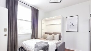 Ein Schlafzimmer mit Bett, einer Bank, einem Kleiderschrank, einem Fenster mit Vorhängen und einem gerahmten Bild an der Wand.