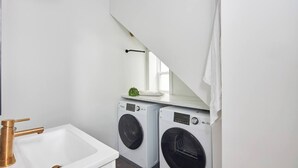 Ein kleiner Waschraum mit Waschmaschine und Trockner, einem Waschbecken mit Goldarmatur sowie einem Fenster über dem Trockner.