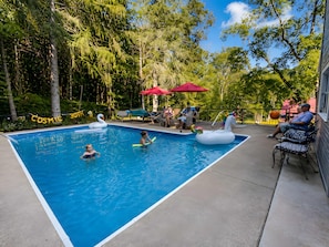A swimming pool with inflatable swan and unicorn floats, surrounded by trees and outdoor seating. - Spacious Family Retreat w/ Pool, Hot Tub, Fire Pit (Ligonier)