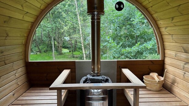 En bastu med träpanel på väggarna, en bänk, en bastuaggregat och utsikt över skogen genom ett stort, välvt fönster.