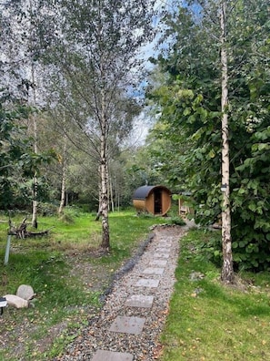 Un chalet isolé, entouré d’arbres, avec un chemin de gravier menant à l’entrée et une petite construction en bois à proximité.
