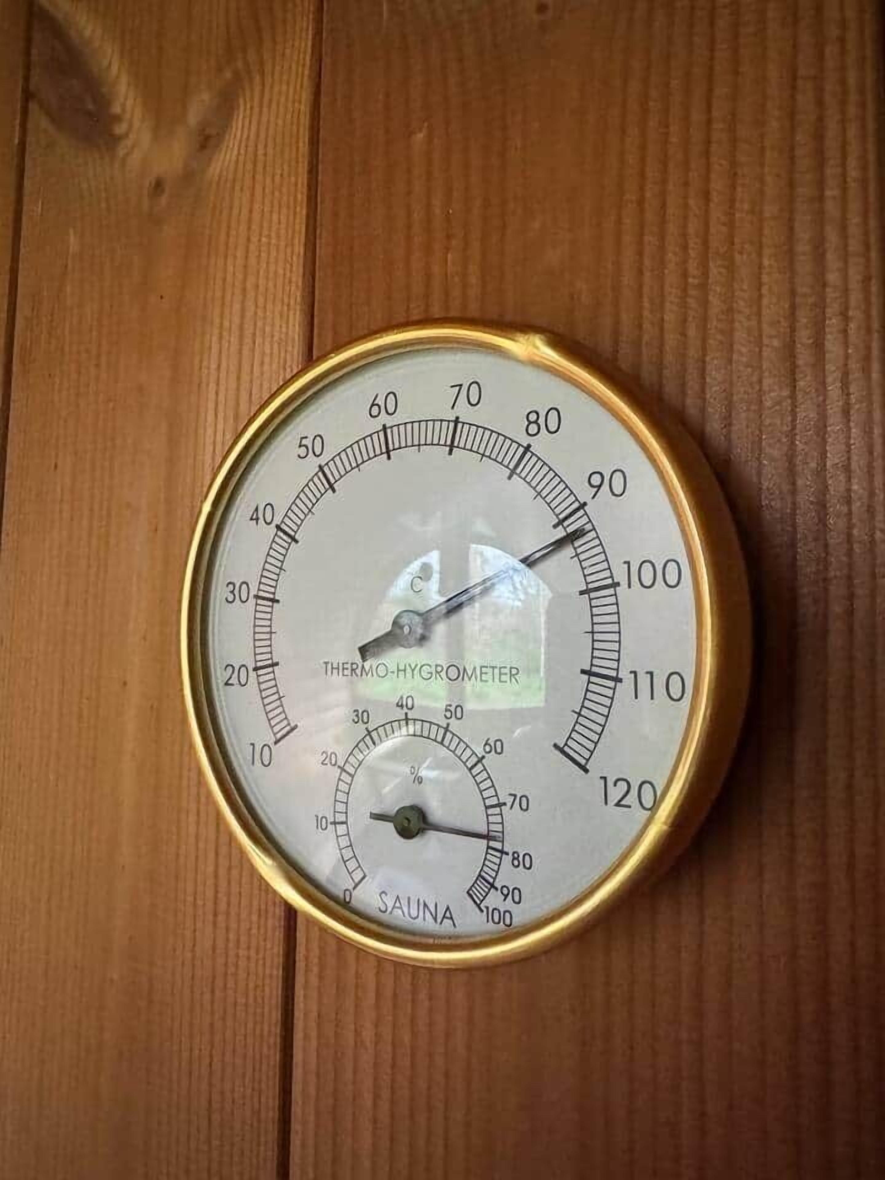 Un thermomètre rond, à cadre doré et de couleur blanche, avec des échelles Celsius et Fahrenheit, et un indicateur de réglage pour sauna.
