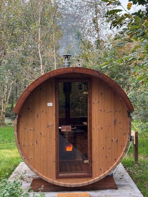 Estructura de sauna de madera con puerta, un banco en el interior y una chimenea que emite humo.