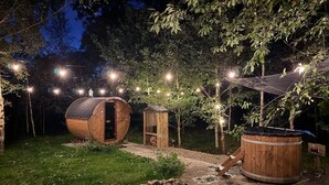 En rustik utomhusmiljö med träkonstruktioner, inklusive en stor byggnad formad som en tunna och ett mindre skjul, omgiven av träd och upplyst av ljusslingor.