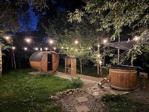 Un entorno rústico al aire libre con construcciones de madera, incluyendo un edificio en forma de barril y un cobertizo más pequeño, rodeado de árboles e iluminado con luces colgantes.