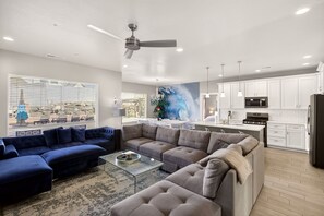 A modern living room with a blue sectional sofa, a grey sectional sofa, and a glass coffee table. There is a kitchen area with white cabinets and a black refrigerator.