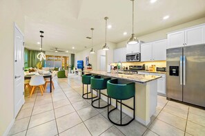 A modern kitchen with a central island, bar stools, a stainless steel refrigerator, and a dining area with a green chair. - New Resort Home, Pool and Spa, Game Room (Davenport)