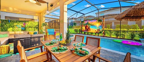 Zona de comedor junto a la piscina con una mesa preparada para cuatro personas, rodeada de sillas de mimbre, un ventilador de techo y vistas a un mural con temática tropical.