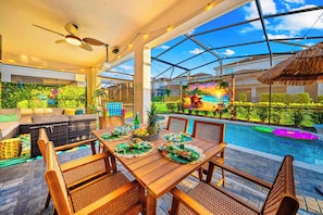 A poolside dining area with a table set for four, surrounded by wicker chairs, a ceiling fan, and a view of a tropical-themed mural. - New Resort Home, Pool and Spa, Game Room (Davenport)