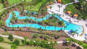 Luftaufnahme eines Resorts mit einem gewundenen Pool, Palmen und einem Strandbereich mit Liegestühlen.