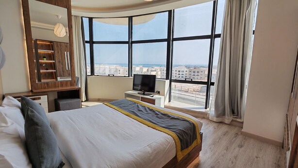 Ein Hotelzimmer mit einem großen Bett, einem Fernseher, einem Schreibtisch und Blick auf die Stadt durch große Fenster.