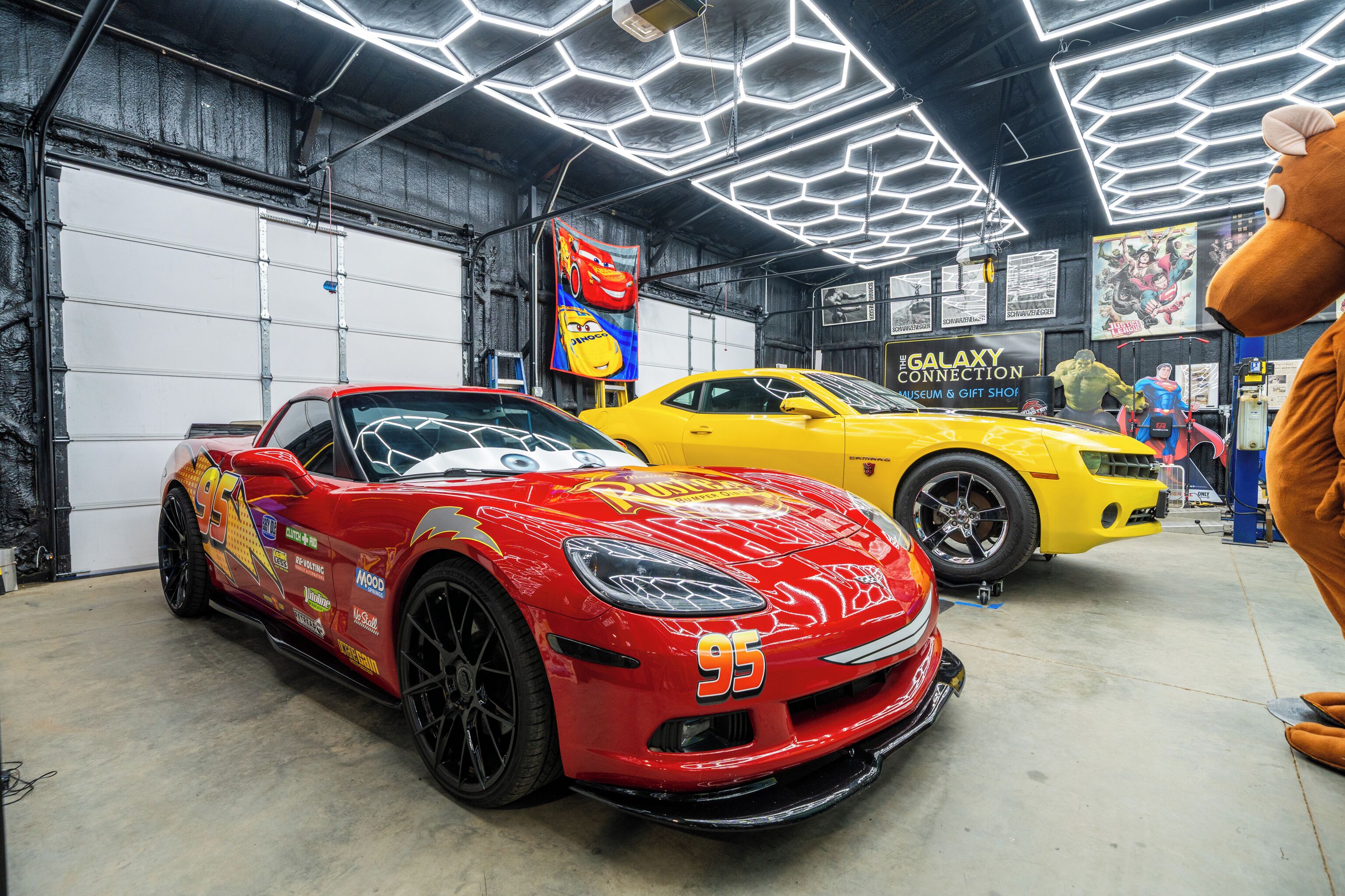 A red sports car and a yellow sports car in a garage with hexagonal lighting and a 'Galaxy Connection' sign.