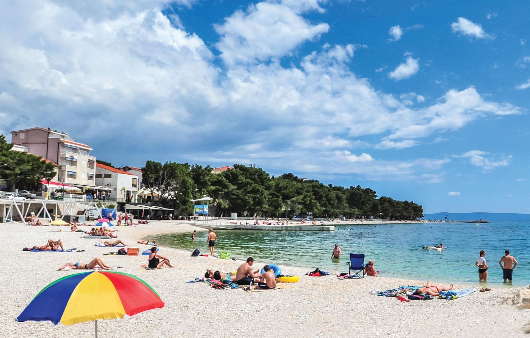 A beach with people sunbathing and swimming, a colorful umbrella, and buildings in the background.