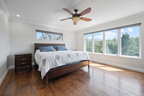 Une chambre avec un plancher en bois, un lit avec une tĂŞte de lit, une table de chevet, un ventilateur de plafond et de grandes fenĂŞtres.