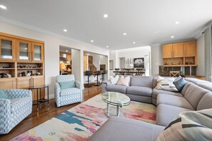A spacious living room with a sectional sofa, a glass coffee table, and patterned armchairs. There are wooden cabinets with glass doors and a kitchen area in the background.