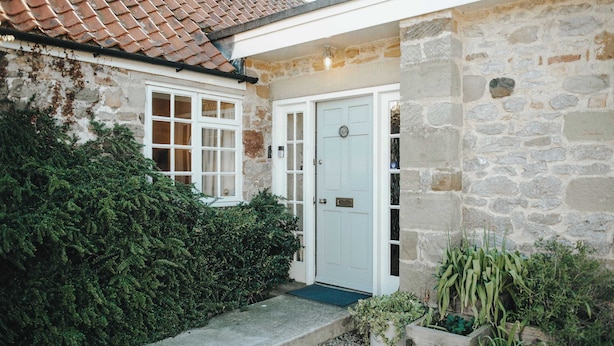 A stone house with a white door, a window, and a small garden area with plants.