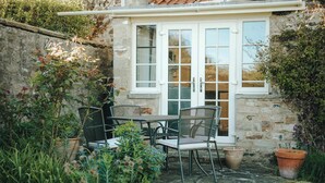 A stone building with a white door and windows, a small patio with a table and chairs, and potted plants.