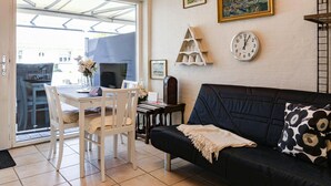 A living room with a black sofa, a white dining table, and chairs. There is a clock on the wall and a framed picture above the sofa.