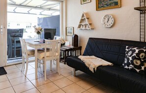 A living room with a black sofa, a white dining table, and chairs. There is a clock on the wall and a framed picture above the sofa.