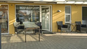An outdoor seating area with a glass table and wicker chairs, a yellow building facade, and a clear view of the street.
