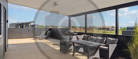 A covered outdoor patio with a wooden deck, a seating area with a sofa and chairs, and a grill.