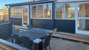 Un patio moderno al aire libre con mesa y sillas, una terraza de madera y una estructura grande con ventanales.